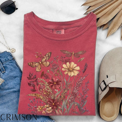 May include: Crimson-colored t-shirt featuring a floral and insect design. The graphic includes a butterfly, dragonfly, and various flowers in shades of yellow, red, and brown. The shirt is folded, with the word "CRIMSON" visible at the bottom.