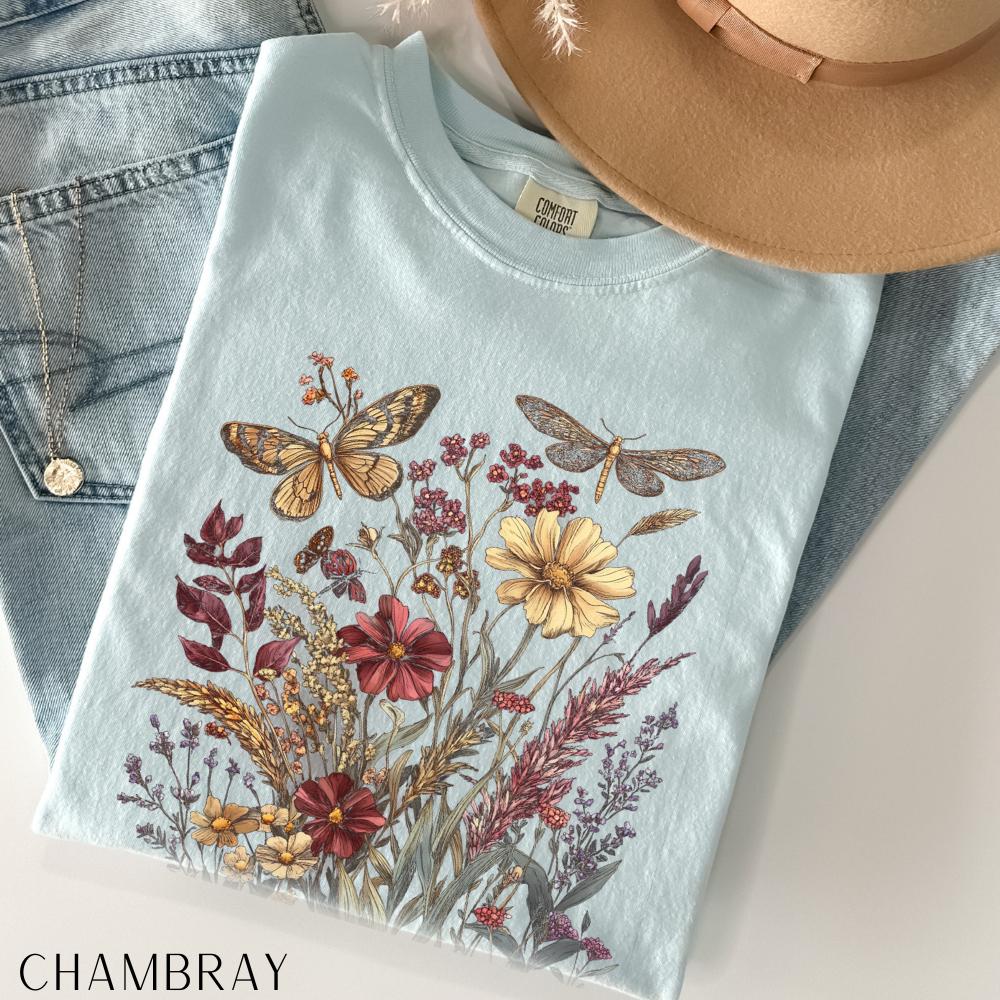 May include: A light blue t-shirt with a floral and butterfly design. The graphic features a variety of flowers in shades of red, yellow, and purple, along with two butterflies. The shirt is paired with blue jeans and a tan hat.