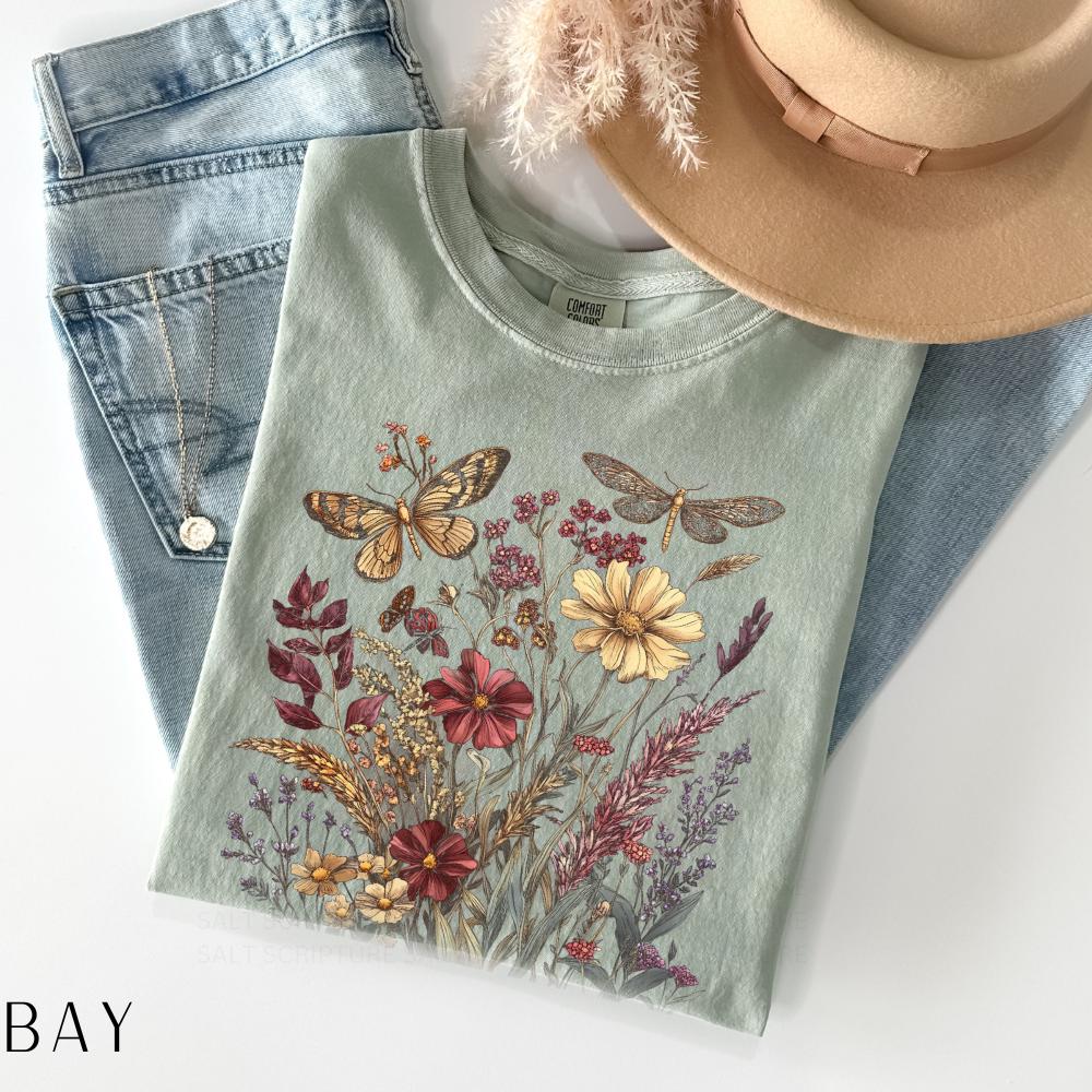 May include: A sage green t-shirt featuring a floral and insect design. The graphic includes butterflies, dragonflies, and various flowers in shades of brown, red, and yellow. The shirt is paired with light blue jeans and a tan hat.