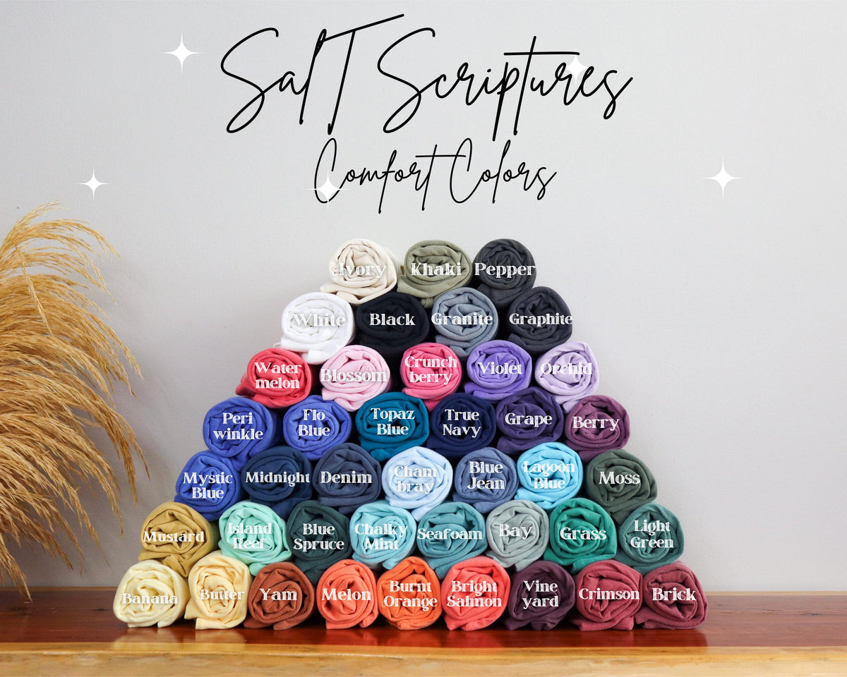 May include: A pyramid of rolled-up t-shirts in various colors, including white, black, and blue. The image features the text "Salt Scriptures Comfort Colors" in a script font. Each shirt is labeled with its color name.