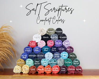 May include: A pyramid of rolled-up t-shirts in various colors, including white, black, and blue. The image features the text "Salt Scriptures Comfort Colors" in a script font. Each shirt is labeled with its color name.