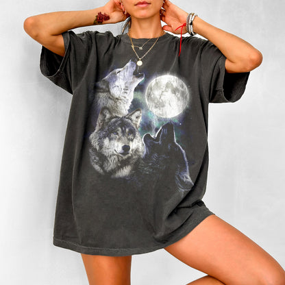 May include: Dark gray t-shirt featuring a graphic of three wolves howling at a full moon. The design includes a detailed illustration of the wolves and the moon against a starry background. The shirt has short sleeves and a relaxed fit.