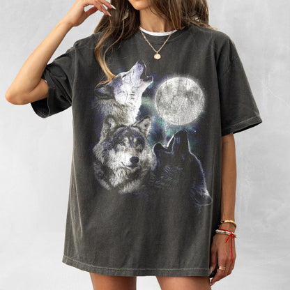 May include: Dark gray t-shirt featuring a graphic of three wolves howling at a full moon. The design includes detailed illustrations of the wolves and a bright, glowing moon against a starry background. The shirt has a vintage, worn look.