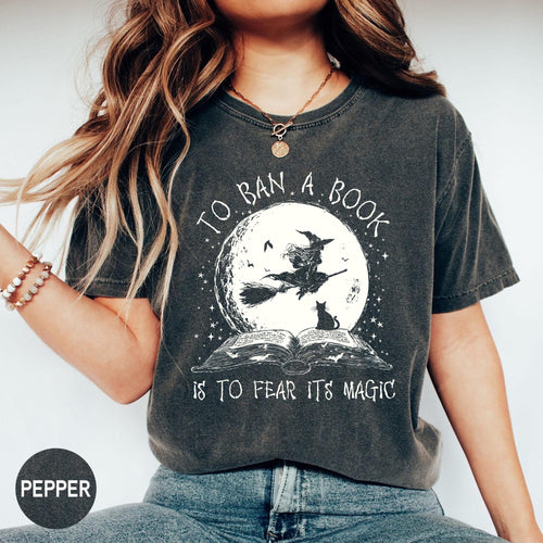 May include: Dark gray t-shirt with the text "TO BAN A BOOK IS TO FEAR ITS MAGIC". The graphic features a witch on a broom in front of a full moon, a black cat, and an open book. The shirt is a Pepper color.
