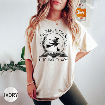 May include: Ivory-colored t-shirt featuring a black graphic of a witch on a broom in front of a moon, above an open book. The text reads "TO BAN A BOOK IS TO FEAR ITS MAGIC."