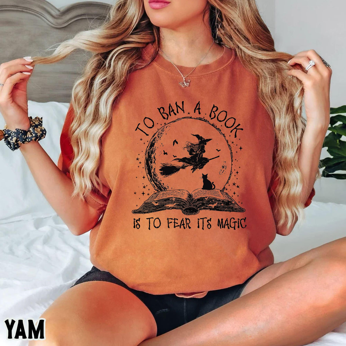 May include: Orange t-shirt with a graphic featuring a witch on a broom in front of a full moon, above an open book. The text reads "TO BAN A BOOK IS TO FEAR ITS MAGIC". The shirt is a comfortable, casual style.