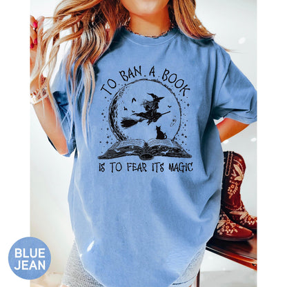 May include: A light blue t-shirt with a graphic featuring a witch on a broom in front of a full moon, a black cat, and the text "TO BAN A BOOK IS TO FEAR ITS MAGIC."