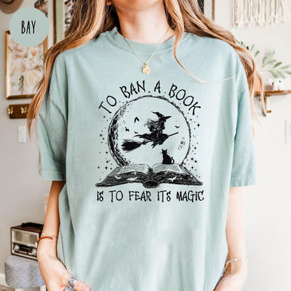 May include: A light blue t-shirt with a graphic featuring a witch on a broom in front of a full moon, a black cat, and an open book. The text reads "TO BAN A BOOK IS TO FEAR ITS MAGIC."