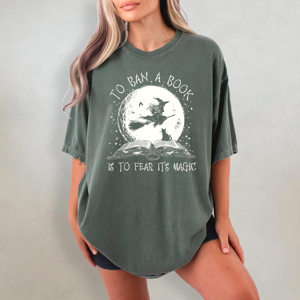 May include: Dark green t-shirt with a graphic featuring a witch on a broom in front of a full moon, a black cat, and an open book. The text reads "TO BAN A BOOK IS TO FEAR ITS MAGIC."