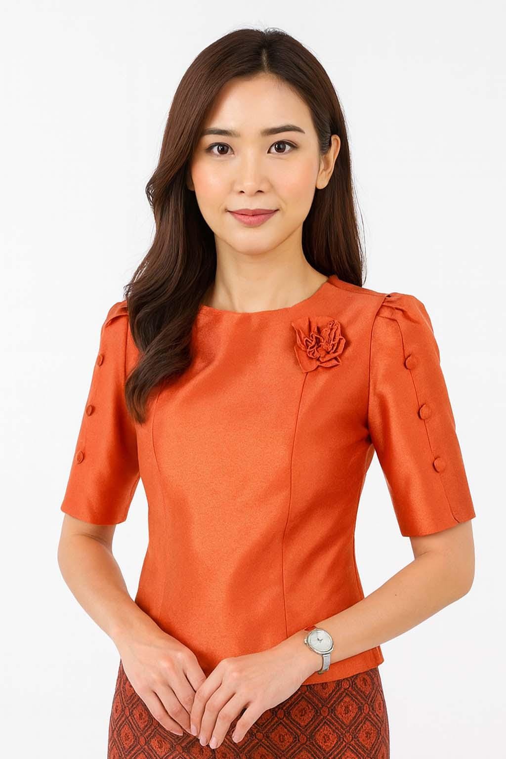 May include: A vibrant orange short-sleeved blouse featuring a round neckline, a decorative fabric flower accent on the left chest, and three matching buttons along the outer edge of each sleeve. The fabric has a subtle sheen and a structured fit.