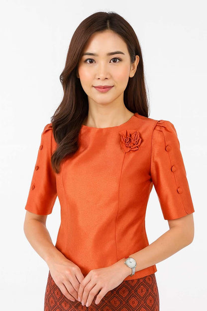 May include: A vibrant orange short-sleeved blouse featuring a round neckline, a decorative fabric flower accent on the left chest, and three matching buttons along the outer edge of each sleeve. The fabric has a subtle sheen and a structured fit.