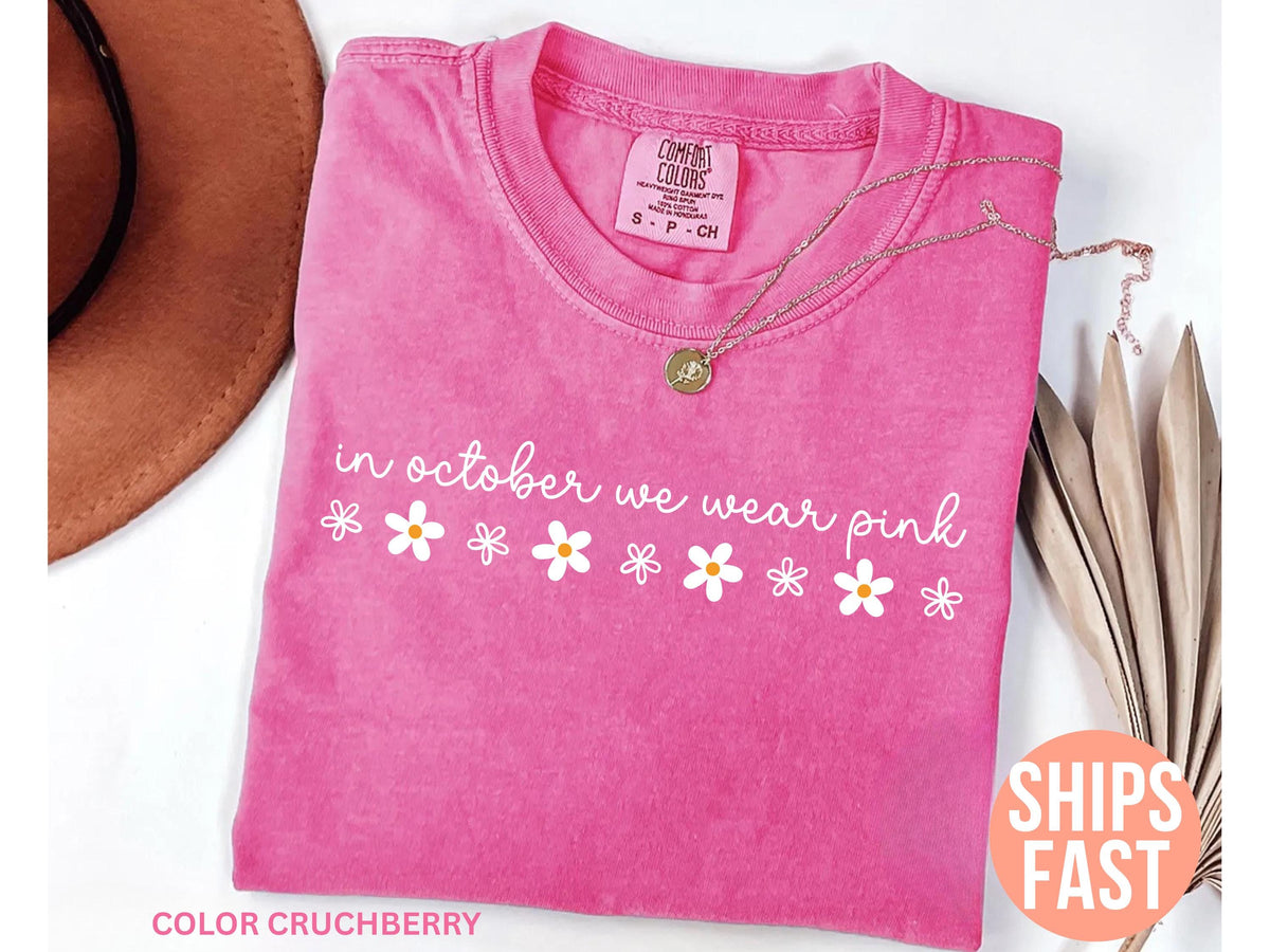 May include: A pink t-shirt with the text "in october we wear pink" and daisy graphics. A gold necklace and a brown hat are also in the image. The shirt is a Comfort Colors brand.