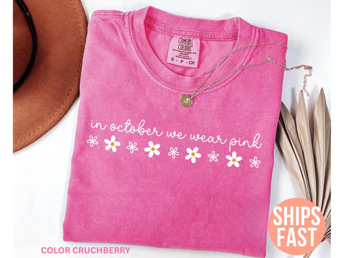 May include: A pink t-shirt with the text "in october we wear pink" and daisy graphics. A gold necklace and a brown hat are also in the image. The shirt is a Comfort Colors brand.
