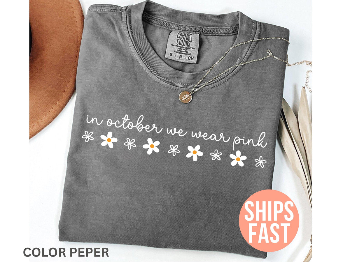 May include: A gray t-shirt with the text "in october we wear pink" in white script and daisy graphics. A gold necklace with a pendant is on the shirt. The shirt has a crew neck and short sleeves.
