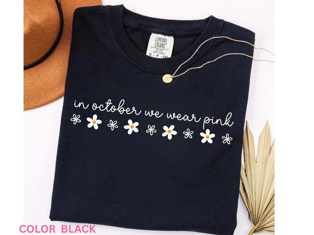 May include: Black t-shirt with the phrase "in october we wear pink" in white script and daisy graphics. A gold necklace and a brown hat are visible in the background. The shirt is a Comfort Colors brand.