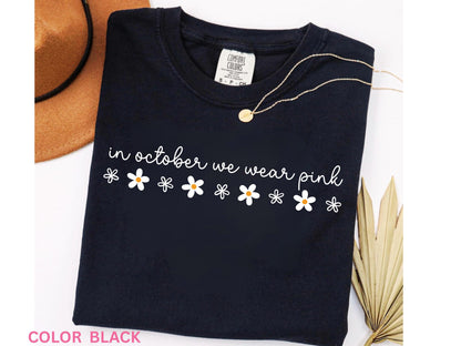 May include: Black t-shirt with the phrase "in october we wear pink" in white script and daisy graphics. A gold necklace and a brown hat are visible in the background. The shirt is a Comfort Colors brand.
