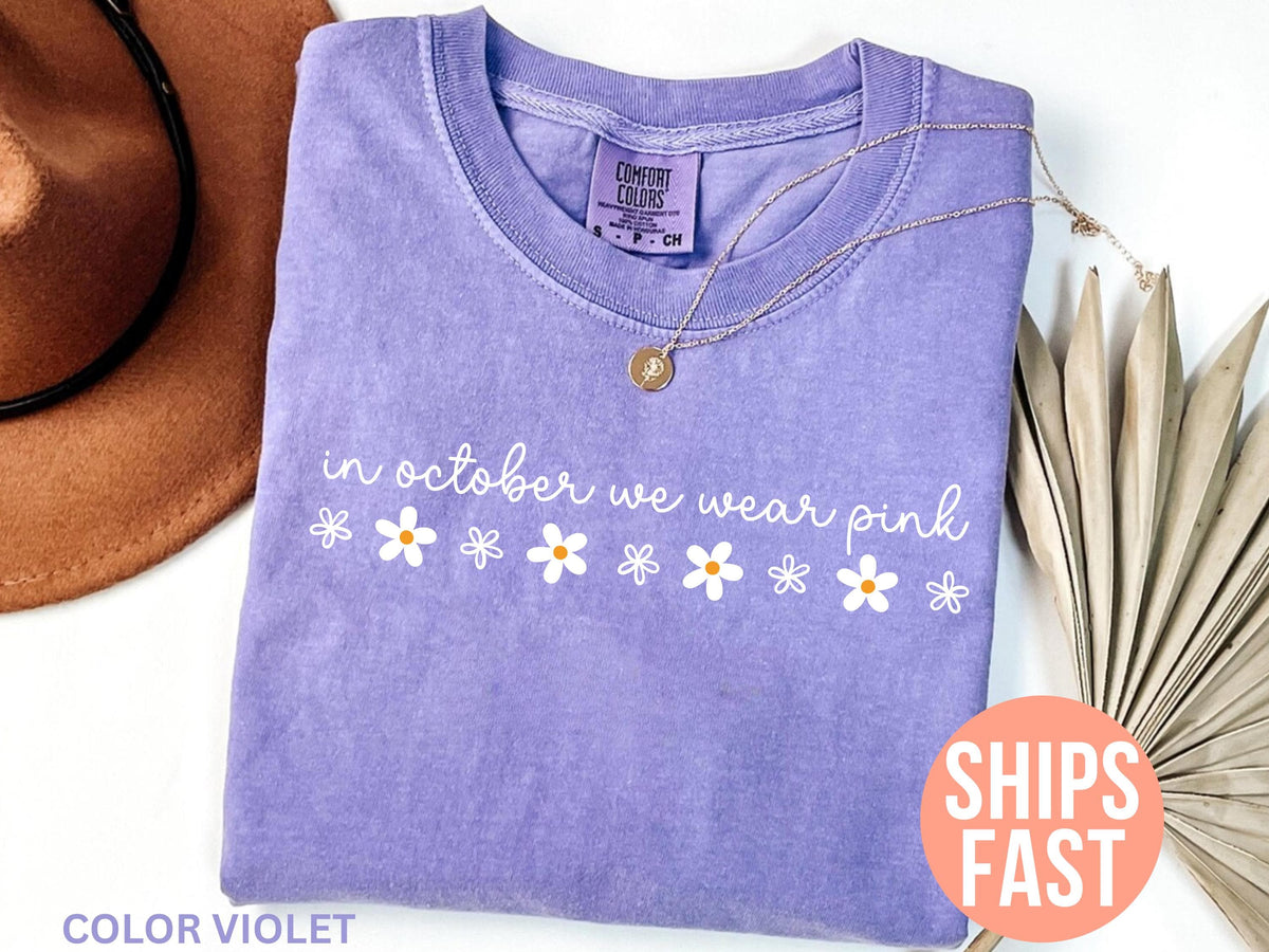 May include: A lavender Comfort Colors t-shirt with the phrase "in october we wear pink" and daisy graphics. A gold necklace and a brown hat are next to the shirt. The shirt is color violet.