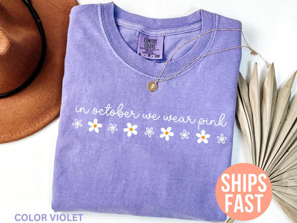 May include: A lavender Comfort Colors t-shirt with the phrase "in october we wear pink" and daisy graphics. A gold necklace and a brown hat are next to the shirt. The shirt is color violet.