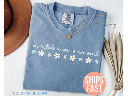 May include: A blue jean colored t-shirt with the text "in october we wear pink" and daisy graphics. A gold necklace is draped across the shirt. The shirt is folded and displayed with a brown hat and dried plant.
