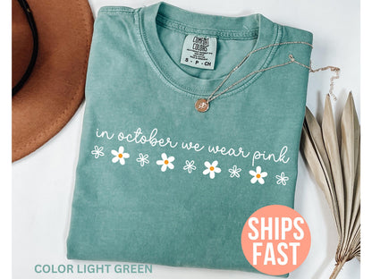 May include: Light green t-shirt with the text "in october we wear pink" and daisy graphics. A silver necklace with a pendant is on the shirt. The shirt is folded and displayed with a brown hat and dried plants.