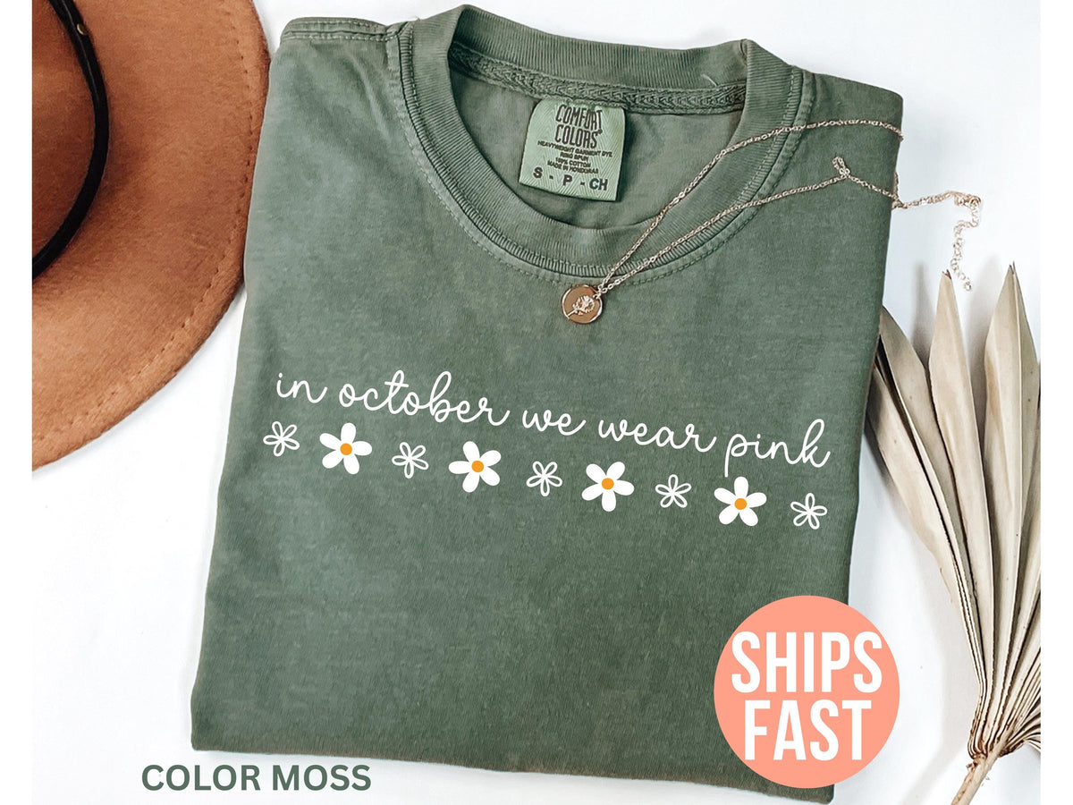 May include: Olive green t-shirt with the text "in october we wear pink" in white script and daisy graphics. A gold necklace with a pendant is draped on the shirt. The shirt is labeled "Color Moss".