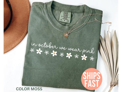 May include: Olive green t-shirt with the text "in october we wear pink" in white script and daisy graphics. A gold necklace with a pendant is draped on the shirt. The shirt is labeled "Color Moss".