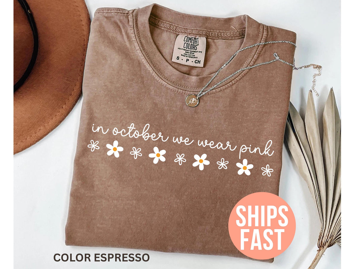 May include: Espresso-colored t-shirt with the text "in october we wear pink" in white script, adorned with daisy graphics. A silver necklace with a pendant rests on the shirt. The shirt has a Comfort Colors tag.