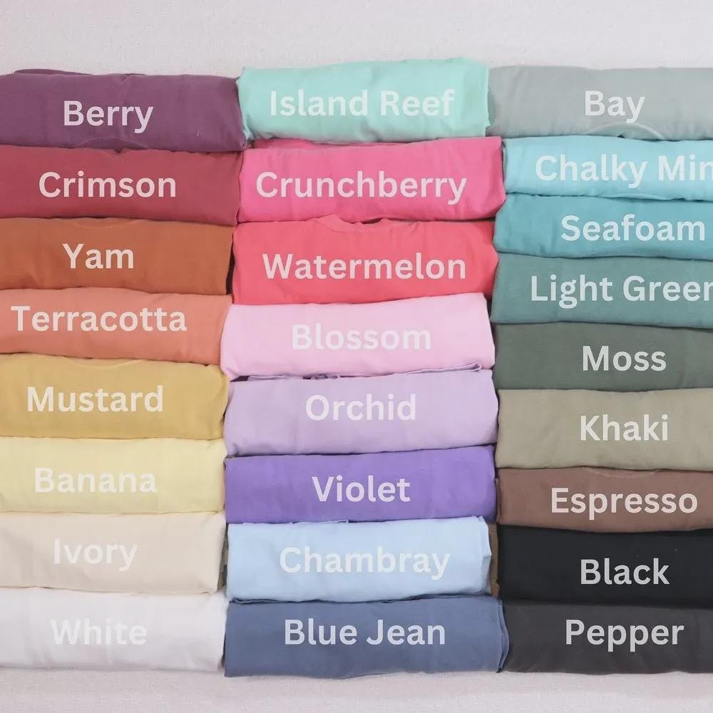 May include: A collection of folded t-shirts in various colors, including Berry, Crimson, Yam, Terracotta, Mustard, Banana, Ivory, White, Island Reef, Crunchberry, Watermelon, Blossom, Orchid, Violet, Chambray, and Blue Jean. Each shirt has a color name printed on it.