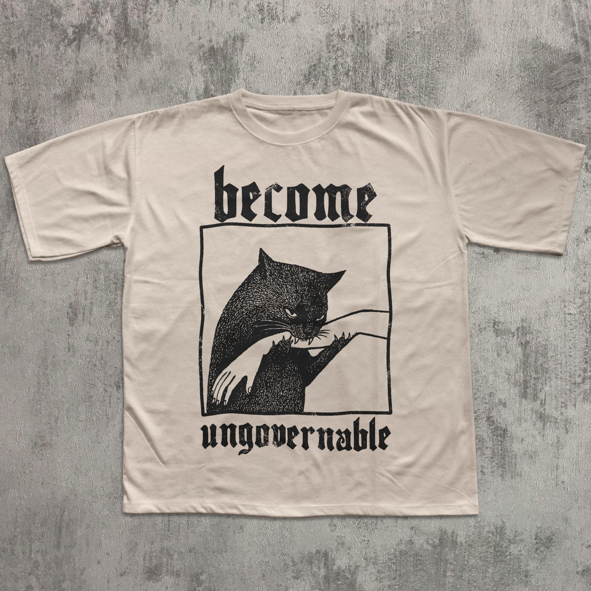May include: Cream-colored t-shirt featuring a black graphic of a cat biting a hand, framed within a rectangle. The words "become" and "ungovernable" are printed above and below the graphic in a gothic font.