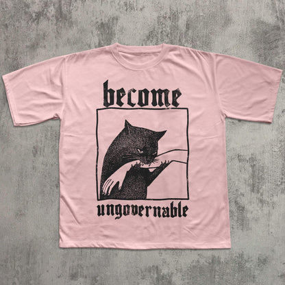 May include: A light pink t-shirt featuring a black graphic of a cat biting a hand, framed within a black rectangle. The words "become" and "ungovernable" are printed above and below the graphic in a gothic font.