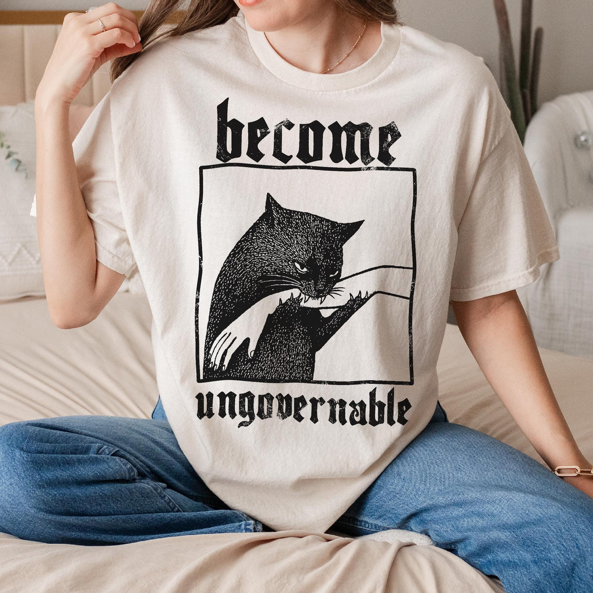 May include: Cream-colored t-shirt featuring a black cat graphic within a black rectangular frame. The text "become" is above the cat, and "ungovernable" is below. The cat graphic shows a larger cat and a smaller cat with a hand reaching out.
