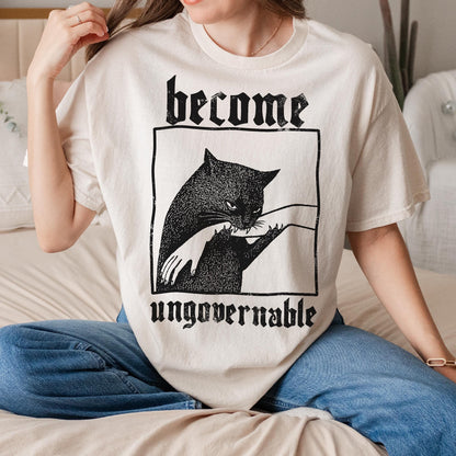 May include: Cream-colored t-shirt featuring a black cat graphic within a black rectangular frame. The text "become" is above the cat, and "ungovernable" is below. The cat graphic shows a larger cat and a smaller cat with a hand reaching out.
