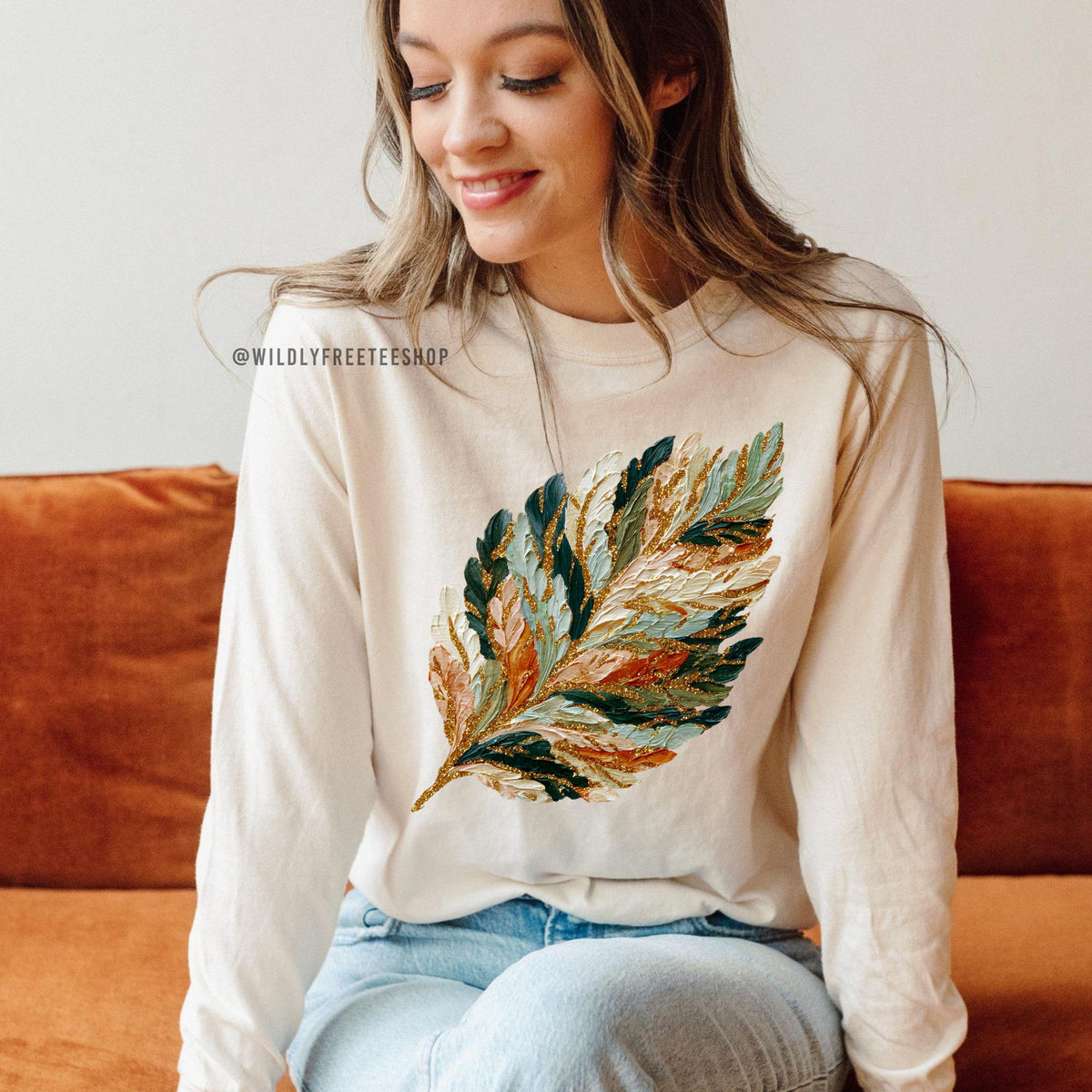 May include: Cream-colored long-sleeve shirt featuring a large, colorful feather design. The feather is detailed with shades of green, blue, orange, and gold. The shirt has the text "@WILDLYFREETEESHOP" printed on it.