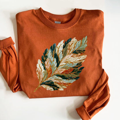 May include: Rust-colored long-sleeve sweatshirt featuring a large, textured leaf design. The leaf is painted with a variety of colors, including white, green, gold, and peach, creating a unique and artistic look. The sleeves are cuffed.