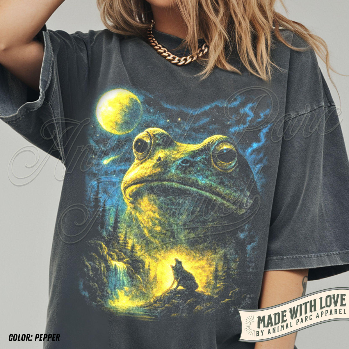 May include: A charcoal gray t-shirt featuring a graphic print of a large frog head above a landscape with a wolf howling at a bright yellow moon. The design includes blue and yellow highlights, a waterfall, and pine trees. The text reads Animal Parc.