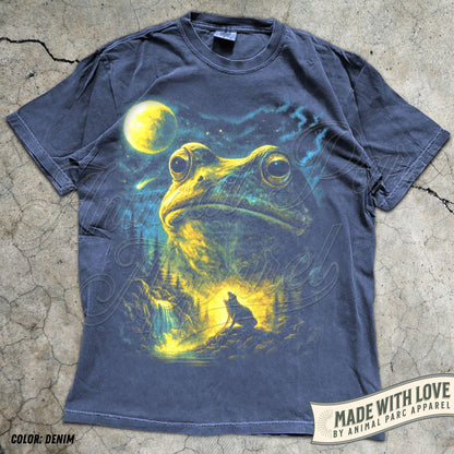 May include: A charcoal gray short-sleeve t-shirt featuring a large graphic print of a giant frog in a night landscape. The scene includes a bright yellow moon, a starry sky, a waterfall, and a small wolf howling at the light in the forest.