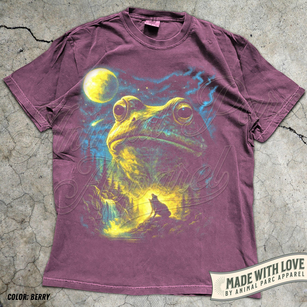May include: A berry-colored short-sleeve t-shirt featuring a graphic print of a large frog looking up at a yellow moon in a starry night sky. Below the frog, a small wolf silhouette stands on a rocky landscape near a waterfall. The shirt has a crew neckline.