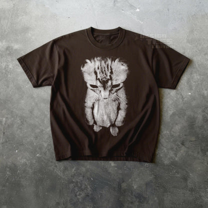 May include: A brown t-shirt featuring a distressed, grayscale image of a sad-looking cat. The cat's face and body are depicted in a stylized, almost sketch-like manner, with a focus on its downturned eyes and posture.