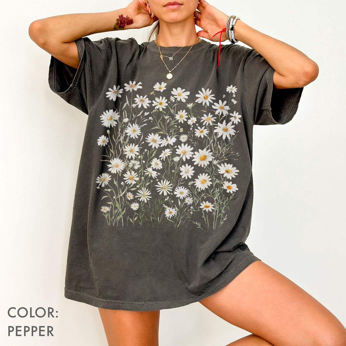 May include: Pepper-colored t-shirt featuring a floral design of white daisies with yellow centers. The graphic is printed on the front of the shirt. The shirt has short sleeves and a relaxed fit.