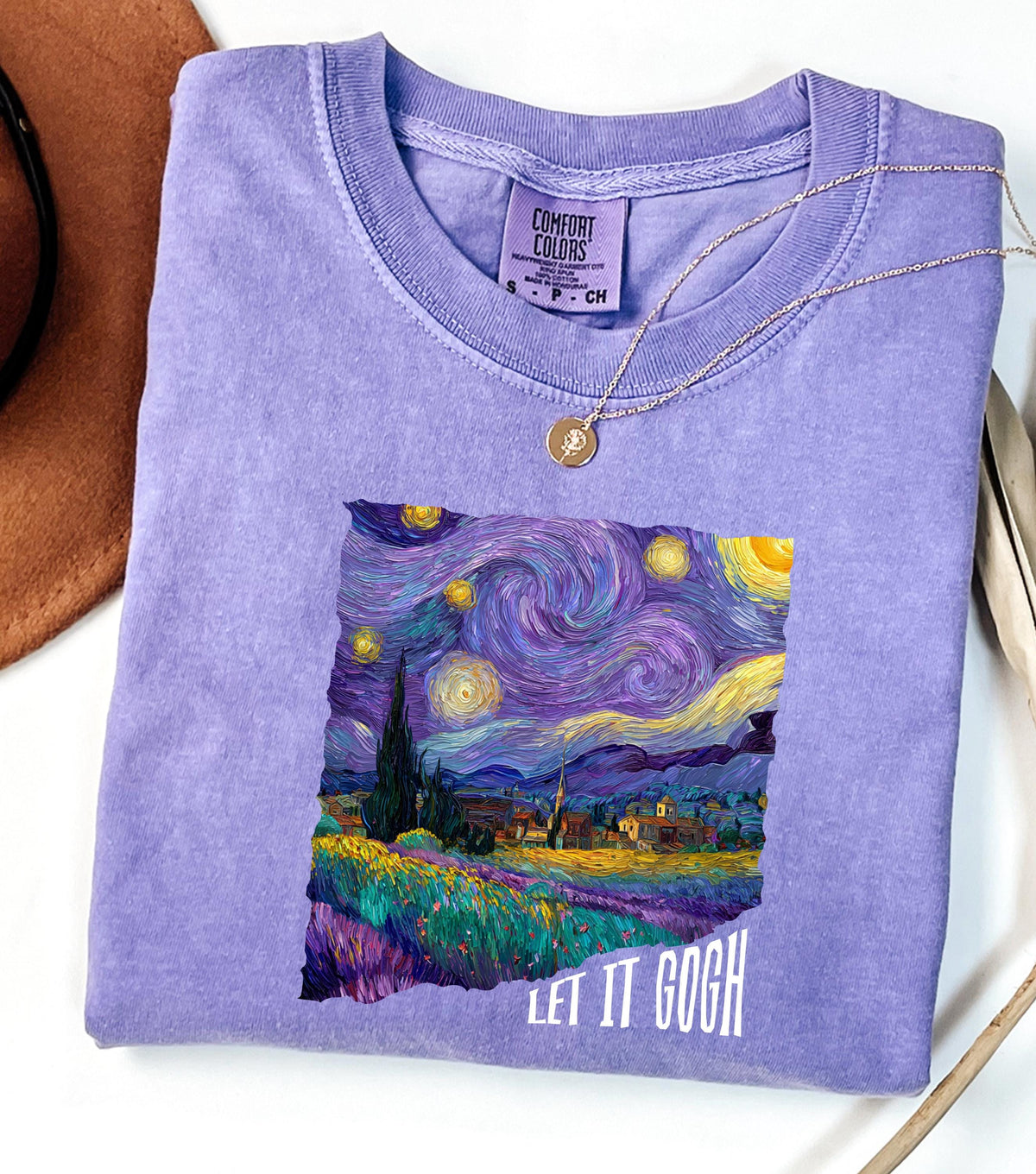 May include: A lilac-colored Comfort Colors t-shirt featuring a print of Van Gogh's "Starry Night" painting. The artwork includes swirling blues, purples, and yellows, with the text "LET IT GOGH" in white. A gold necklace rests on the shirt.
