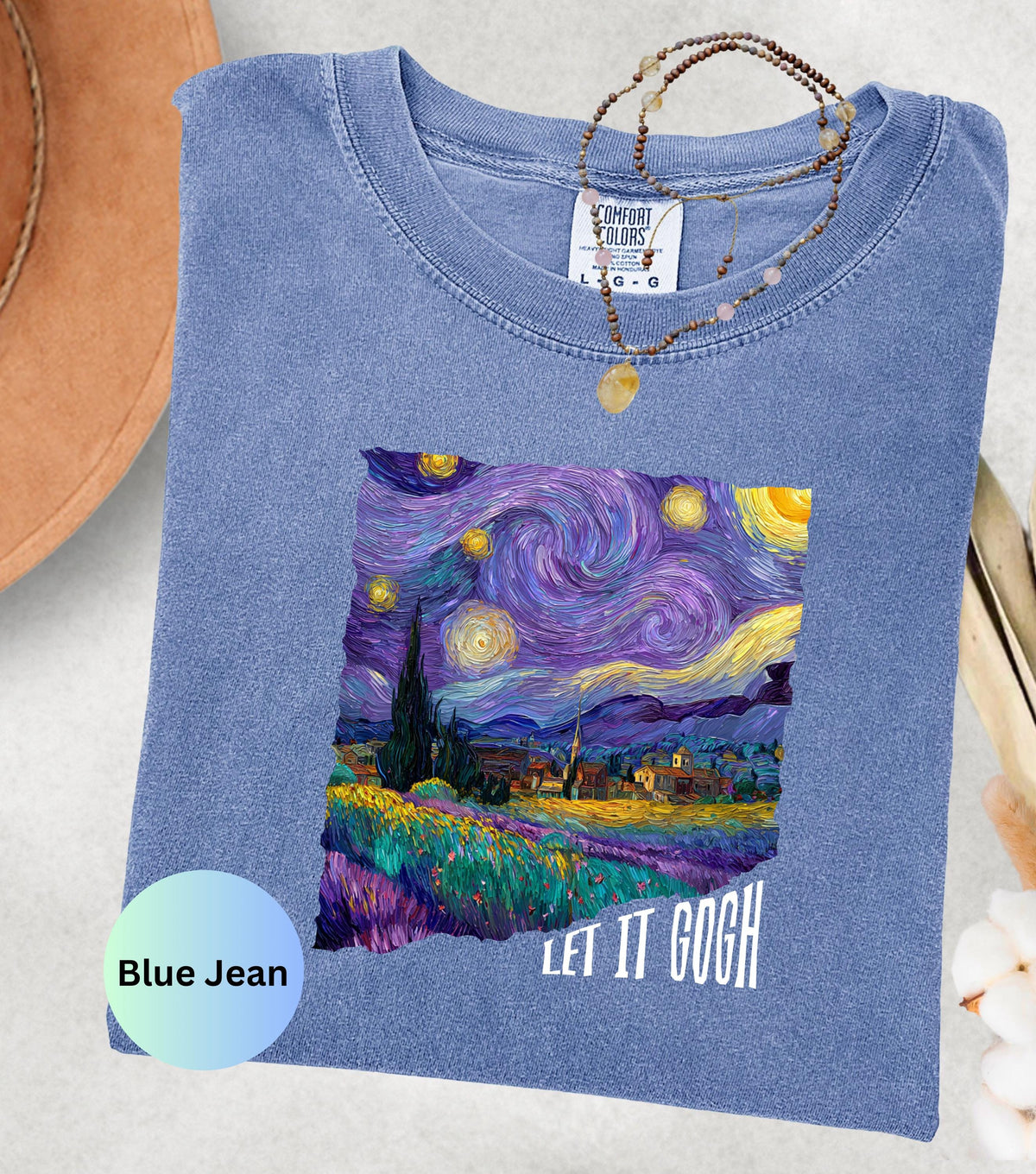 May include: Blue jean colored t-shirt featuring a vibrant print of Van Gogh's "Starry Night" painting. The shirt has the text "LET IT GOGH" in white. A necklace with a pendant rests on the shirt.