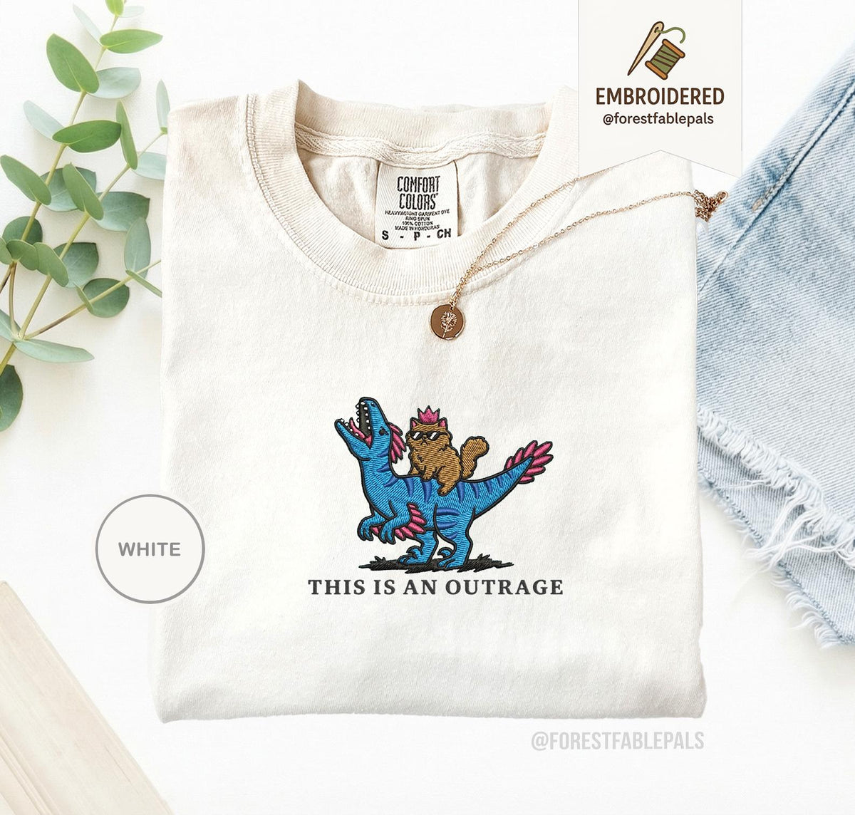 May include: White Comfort Colors t-shirt with an embroidered design. The design features a brown cat wearing sunglasses riding a blue dinosaur with pink accents. The text "THIS IS AN OUTRAGE" is below the design.