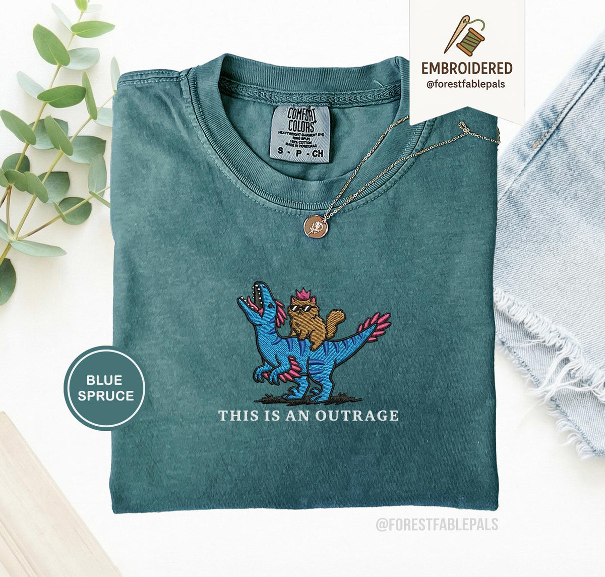 May include: Blue spruce t-shirt with an embroidered design of a cat riding a blue dinosaur. The text "THIS IS AN OUTRAGE" is below the design. The shirt has a crew neck and short sleeves.