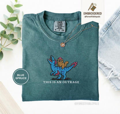 May include: Blue spruce t-shirt with an embroidered design of a cat riding a blue dinosaur. The text "THIS IS AN OUTRAGE" is below the design. The shirt has a crew neck and short sleeves.