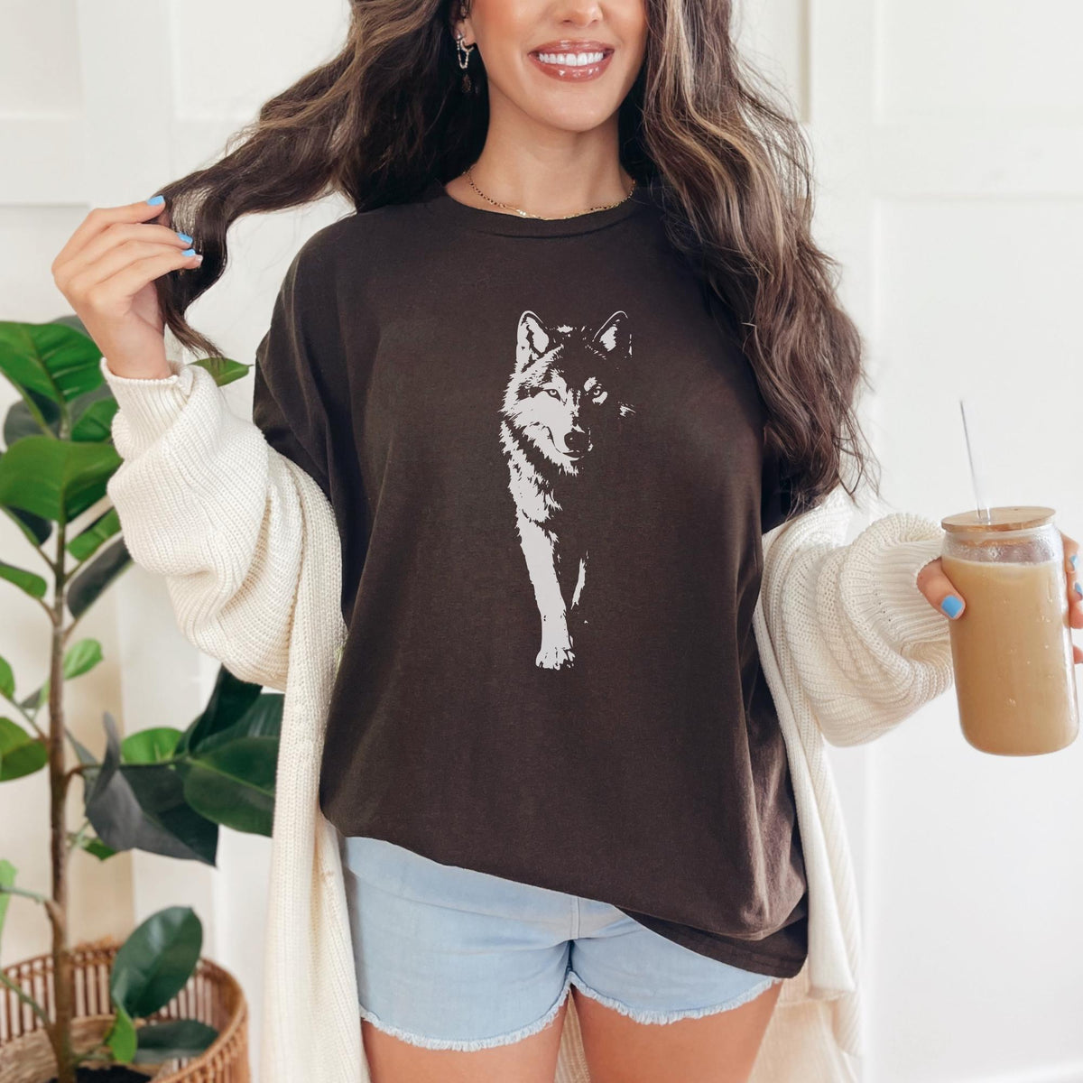 May include: A dark brown t-shirt featuring a white wolf graphic. The shirt is worn with light blue denim shorts and a cream-colored cardigan. A person is holding a glass of iced coffee.