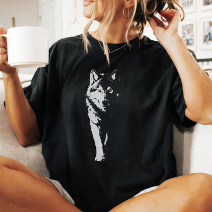 May include: Black t-shirt featuring a white graphic of a wolf. The wolf is depicted in a detailed, artistic style, with a focus on its face and front legs. The shirt is a casual style, suitable for everyday wear.