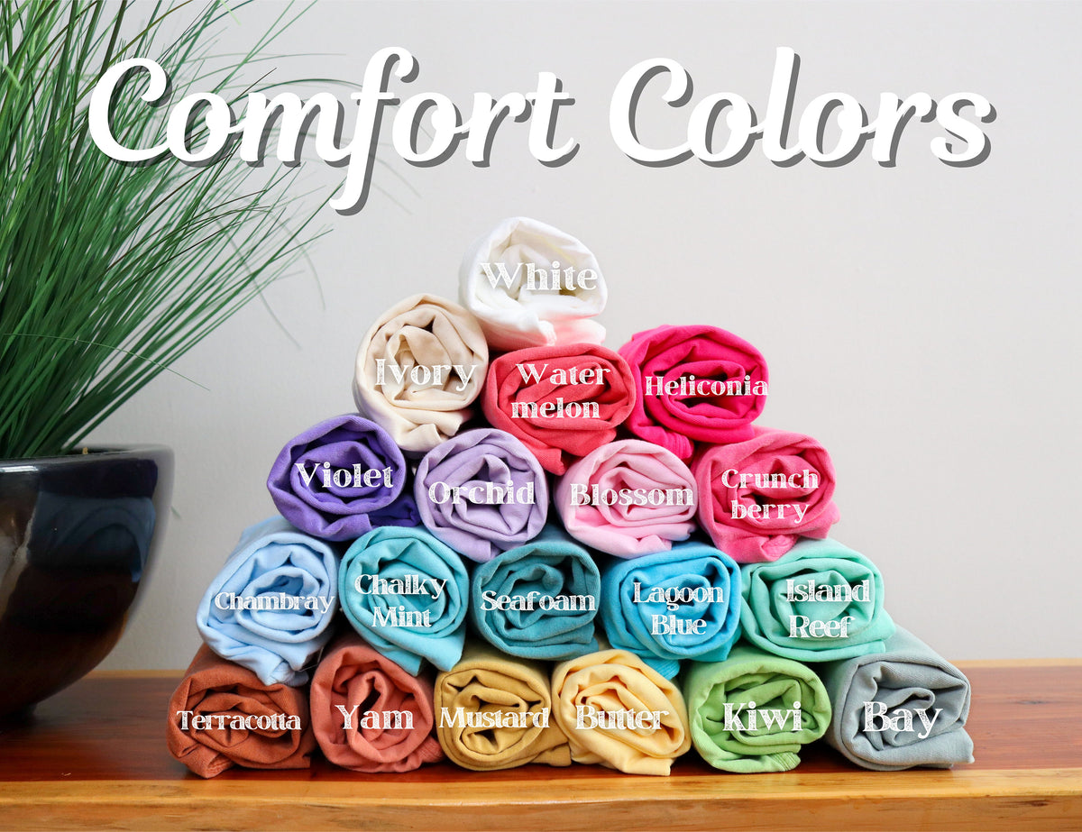 May include: A display of rolled-up t-shirts in various colors, including white, ivory, violet, and lagoon blue. The image features the text "Comfort Colors" in a script font, with each shirt labeled with its color name.