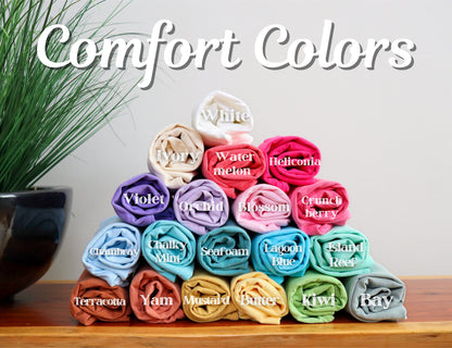May include: A display of rolled-up t-shirts in various colors, including white, ivory, violet, and lagoon blue. The image features the text "Comfort Colors" in a script font, with each shirt labeled with its color name.