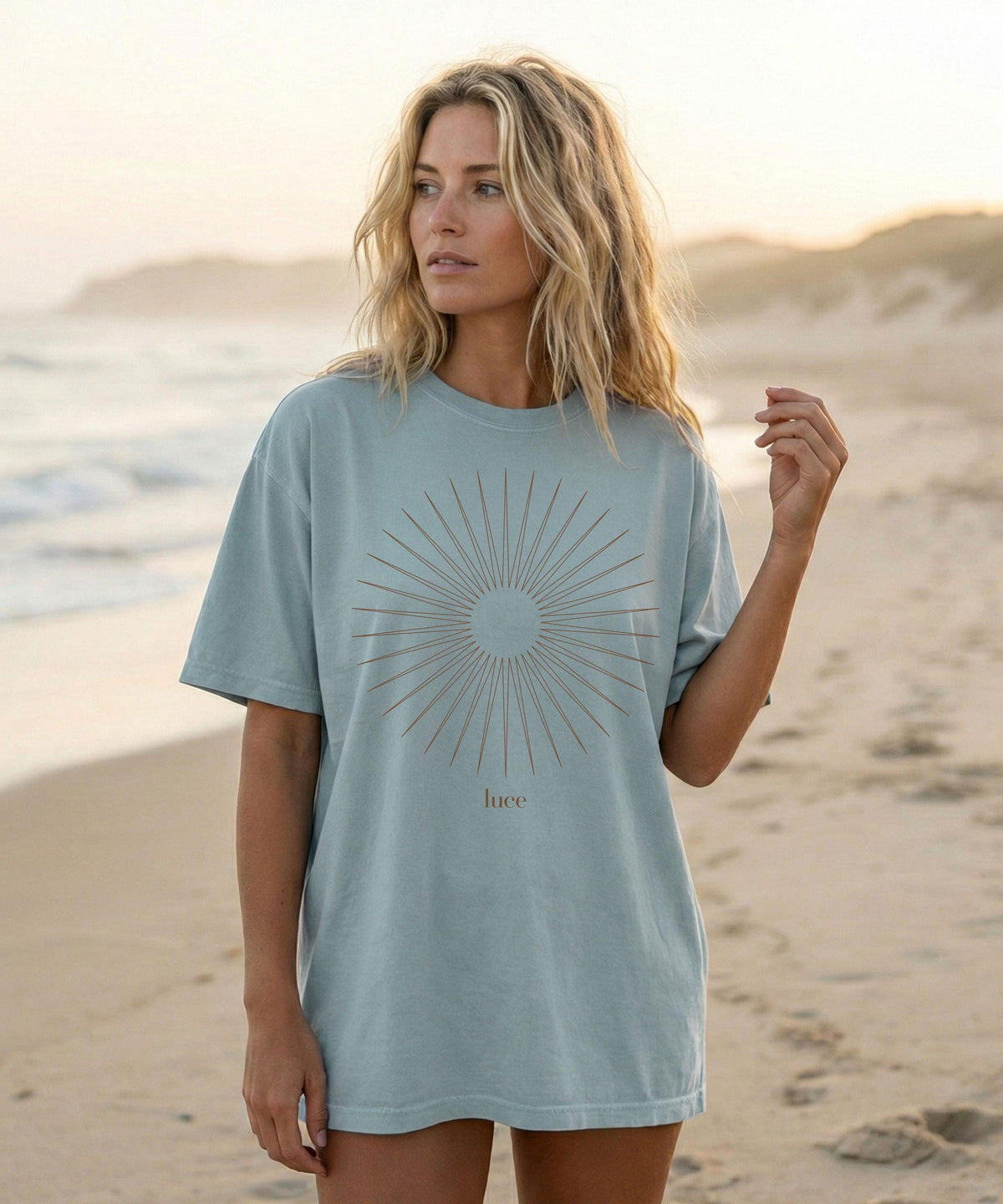 May include: A light blue t-shirt featuring a sunburst design with the word "luce" in a brown font. The shirt is made of a soft material and has short sleeves. The model is standing on a sandy beach.