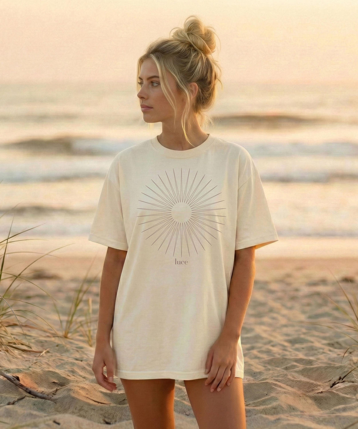 May include: Cream-colored t-shirt featuring a sunburst design with the word "luce" below it. The shirt is worn on a beach with the ocean in the background. The sunburst design is in a dark grey color.
